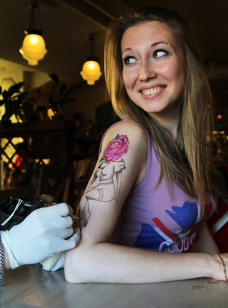 tatuador que muestra el proceso de hacer un tatuaje en la joven hermosa mujer hipster con la mano de pelo rubio. Diseño del tatuaje en la forma pin-up chica con cabeza de flor
 - Foto, Imagen