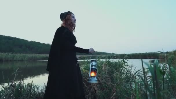 young female dressed in old-fashioned clothes standing near the lake with a lamp - Footage, Video