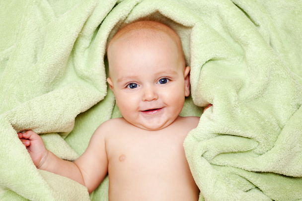 Caucasian baby boy covered with green towel joyfully smiles at c - Zdjęcie, obraz