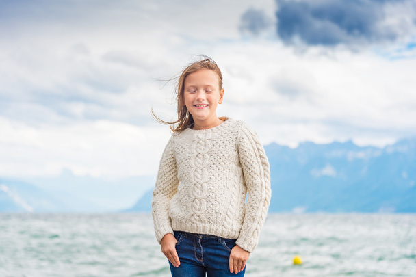 niedliches kleines Mädchen von 8 Jahren, das an einem sehr windigen Tag am See spielt und einen warmen weißen Strickpullover trägt - Foto, Bild