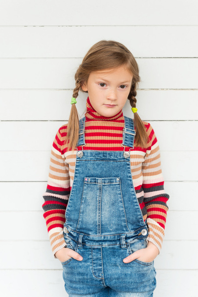 Модный портрет милой маленькой девочки на белом фоне, в полосатом свитере и джинсовом комбинезоне
 - Фото, изображение