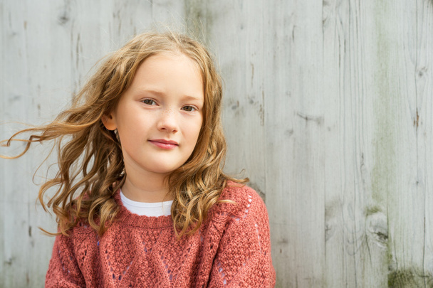Nahaufnahme Porträt eines süßen kleinen Mädchens von 8 Jahren mit lockigem Haar, das Terrakottapullover trägt und vor grauem Holzhintergrund sitzt - Foto, Bild