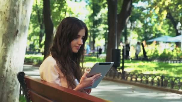 vrouwelijke student zittend op een bankje en met behulp van een tablet in park - Video