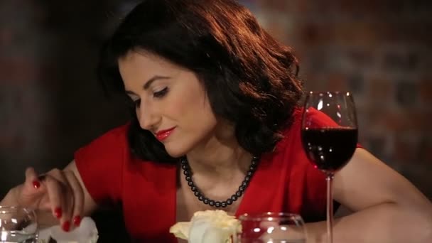 Romantische Brunette in een rode jurk met een roos, kaarsen en wijn glimlachend en flirten - Video