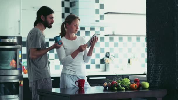 γυναίκα χρησιμοποιώντας ένα δισκίο στέκεται στην κουζίνα με τον άνθρωπο που κατέχει ένα φλυτζάνι - Πλάνα, βίντεο