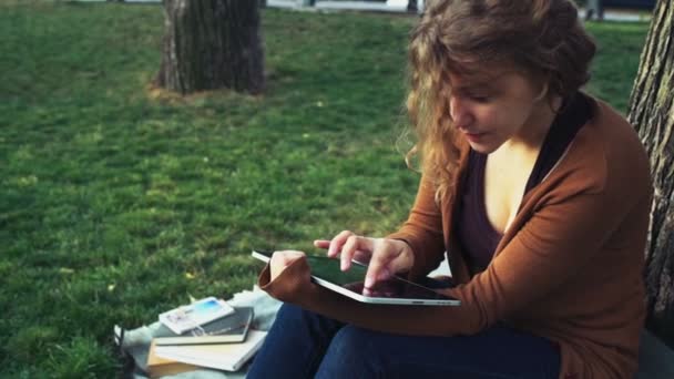 female student sitting under a tree in park and using tablet - Materiał filmowy, wideo