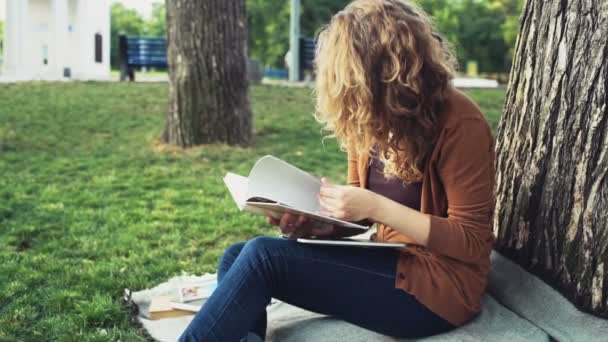 female student flipping the notebook under a tree in park slow motion - Footage, Video