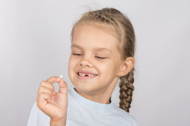 Шестилетняя девочка с улыбкой смотрит на выпавший молочный зуб
 - Фото, изображение