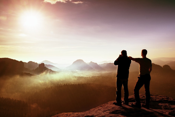Двое друзей. Туристы и фотолюбители фотографируются на скале. Мечтательный туманный пейзаж, голубой туманный восход солнца в красивой долине внизу
 - Фото, изображение