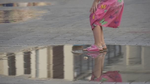 The Little Girl Dressed in a Summer Clothes Plays in the Puddle on Crowded Street. - Video