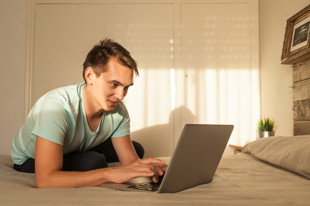 Sorridente giovane uomo biondo utilizzando un computer portatile in camera da letto
. - Foto, immagini