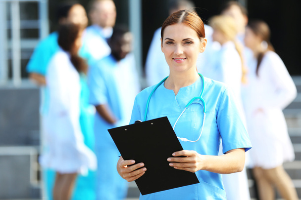 Portrait of woman doctor with clipboard in hands standing against unfocused group of medics - Fotoğraf, Görsel