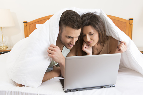 Young couple snuggled under the cover with laptop - 写真・画像