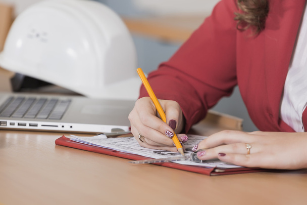 図面前のテーブルに座っている赤いスーツの女性建築家。コンピューターがテーブルに。鉛筆で女子学生は。ディレクター. - 写真・画像