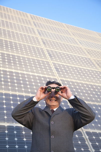 Businessman looking through binoculars in front of solar panels - Photo, Image