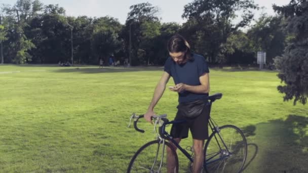 若い男が電話で公園と textes メッセージで彼の自転車のそばに立つ - 映像、動画