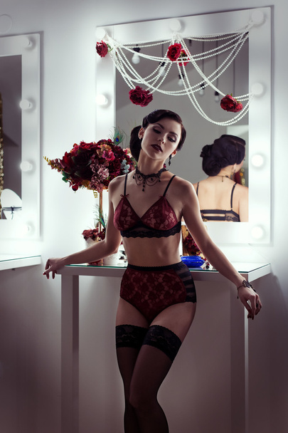 beautiful sexy young woman flaunts front of the mirror in the dressing room in lacy underwear in a retro style with bright make-up and beautiful evening hairstyles , shot in the style of the 20s - Photo, image