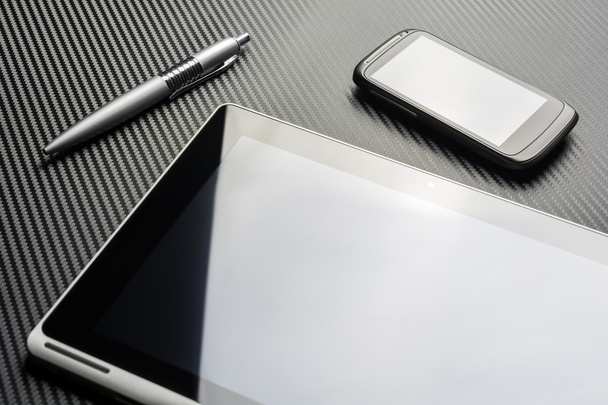 leere Business-Smartphones und ein Stift, der neben einem Tablet mit Reflexion über einem Carbon-Hintergrund liegt - Foto, Bild