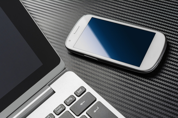 Trabajo de negocios con teléfono inteligente blanco en blanco con reflexión azul que miente derecho a un teclado portátil, todo por encima de una capa de carbono
 - Foto, imagen