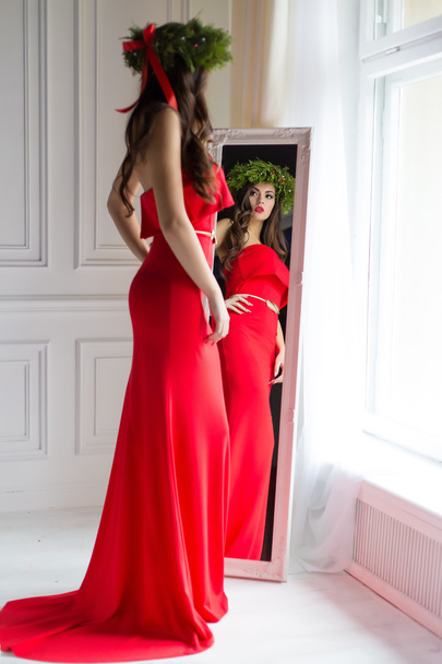 beautiful sexy woman in elegant long evening red dress standing in the mirror next to the window with a Christmas wreath on her head , the image of New Year's Eve - Photo, Image