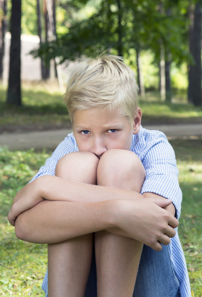 Θλιβερή αγόρι που αγκάλιαζε γόνατά και κάθεται στο πράσινο γρασίδι στην φύση. - Φωτογραφία, εικόνα