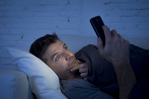 junger Mann in der Schlafcouch zu Hause spät in der Nacht mit intensivem Gesichtsausdruck mit Handy bei schwachem Licht - Foto, Bild