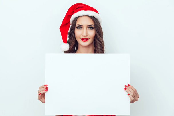 Beautiful girl in the hat of Santa Claus. Hold a sheet of paper. Red manicure. Red lips. - Foto, Imagen