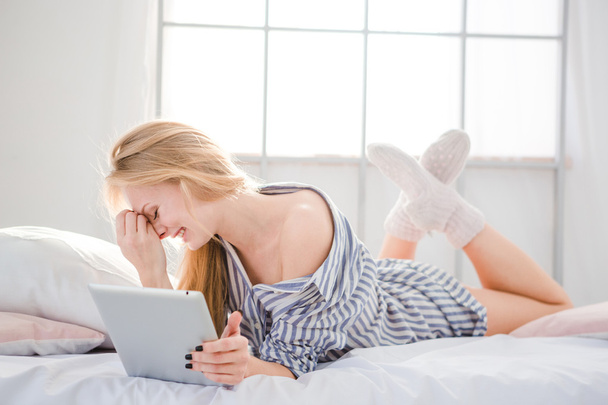 Embarrassed cute woman lying in bed and using tablet - Foto, afbeelding