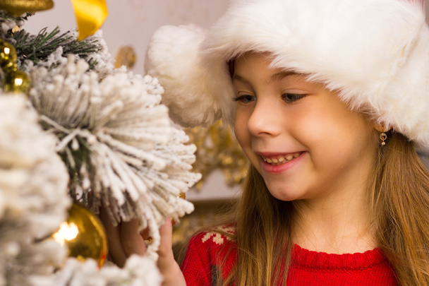 Cute happy girl in red hat hangs decoration on christmas tree - Fotó, kép