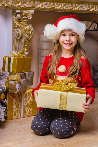 Cute happy girl in red hat holding present near christmas tree - Zdjęcie, obraz