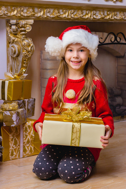 Cute happy girl in red hat holding present near christmas tree - 写真・画像