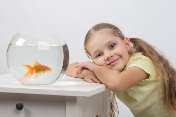 Happy four-year girl put her head in his hands sitting in front of a goldfish - Photo, Image