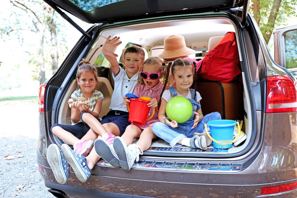 Tres hermosas chicas y un niño se sientan en el maletero de un coche y se ríen
 - Foto, Imagen