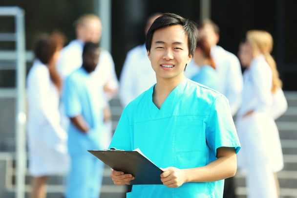 Jovem médico sorridente com prancheta nas mãos em pé contra o grupo de trabalhadores médicos
 - Foto, Imagem