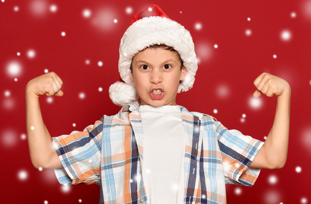 vacaciones de invierno concepto de Navidad - niño en sombrero de santa mostrar músculo
  - Foto, Imagen