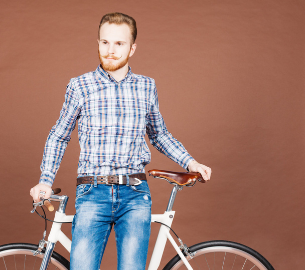 Un joven con bigote y barba está cerca de la moderna bicicleta de accesorios de moda. Jeans y camisa a cuadros estilo hipster. Color tonificado
 - Foto, Imagen