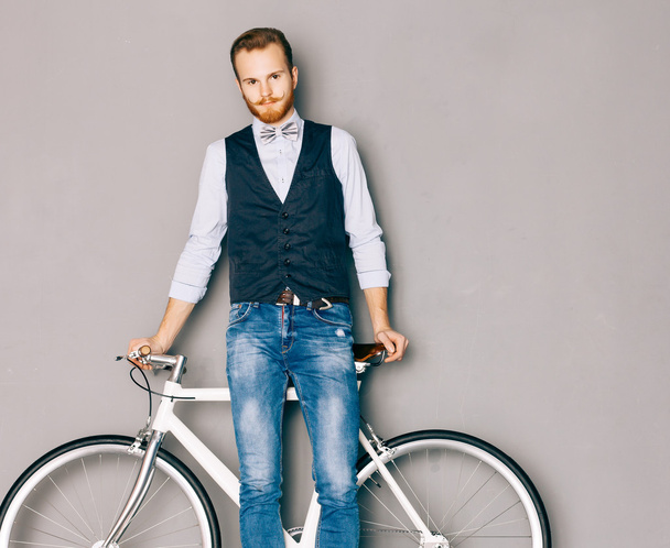 A young man with mustache and beard is near fashionable modern fixgear bicycle. Jeans and shirt, vest and the bow tie hipster style. Gray background. Toned color. Looking at the camera - Photo, Image