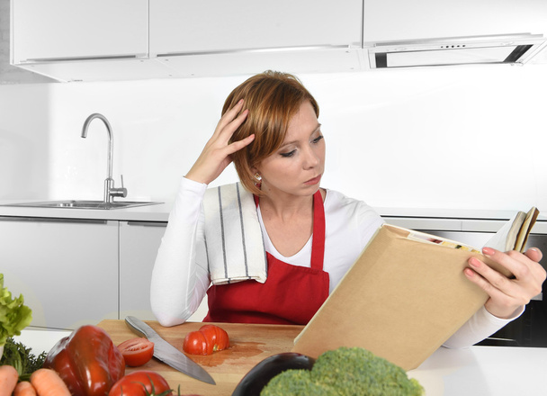 jeune belle maison cuisinière femme en tablier rouge à la cuisine domestique moderne lecture livre de cuisine suivant la recette
 - Photo, image