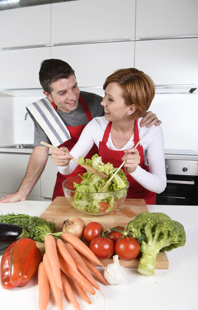 beautiful American couple working at home kitchen in apron mixing vegetable salad smiling happy - Photo, Image