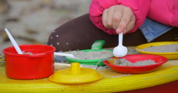 Little Girl's Hands With PLastic Spoon Close View Girl Plays With Colorful Toy Dishes In The Yard She Puts Sand In Toy Cookware Autumn Day Outdoors - Кадри, відео