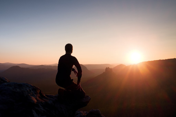Jonge man in zwarte sportkleding zitten op de rand van de klif en kijken naar daybreak aan horizon over mistige vallei  - Foto, afbeelding