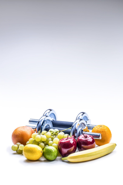 Dumbbells surrounded with healthy fruits measuring tape on a  white background with shadows. - Foto, Imagem
