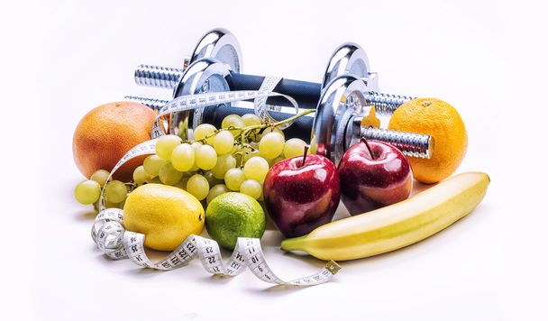 Dumbbells surrounded with healthy fruits measuring tape on a  white background with shadows. - Zdjęcie, obraz