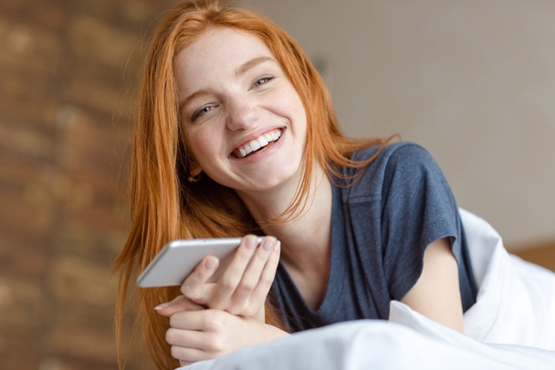 Redhead vrouw liggend op het bed met smartphone - Foto, afbeelding