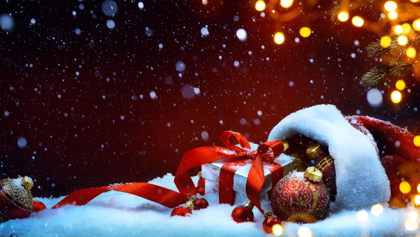Красная сумка Санта-Клауса с рождественскими шарами и подарочной коробкой на снегу
 - Фото, изображение