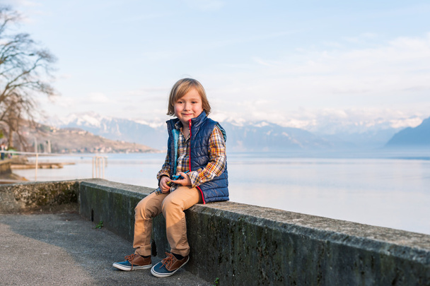 Outdoor portrait of a cute fashion little boy of 4 years old, wearing blue waistcoat, plaid shirt, beige trousers and sneakers - Photo, Image