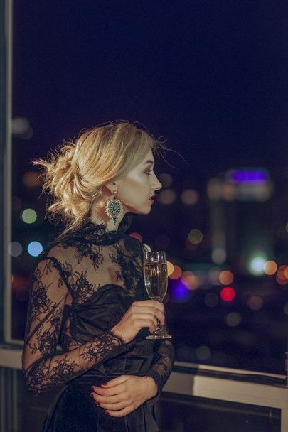 pretty lady with glass of champagne looking through window to night city - Foto, Bild