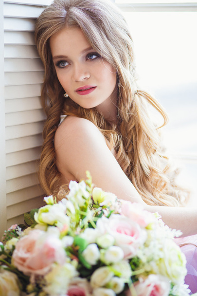 Portrait of pretty young girl in a sweet dress and long blonde hair sitting on a window with flowers composition - Фото, зображення