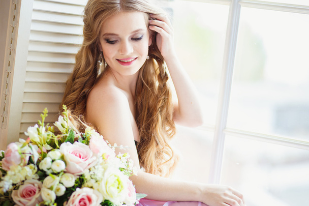 Portrait of pretty young girl in a sweet dress and long blonde hair sitting on a window with flowers composition - Fotó, kép