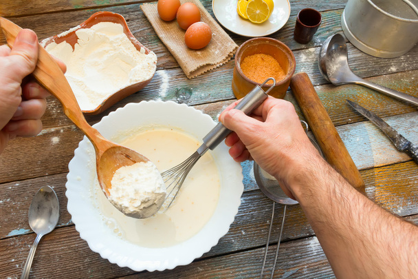The man knead the dough with a whisk. Wheat flour, batter, eggs, a lemon and kitchen utensils on wooden table. Preparation of the dough in a rustic kitchen. - Foto, Imagen